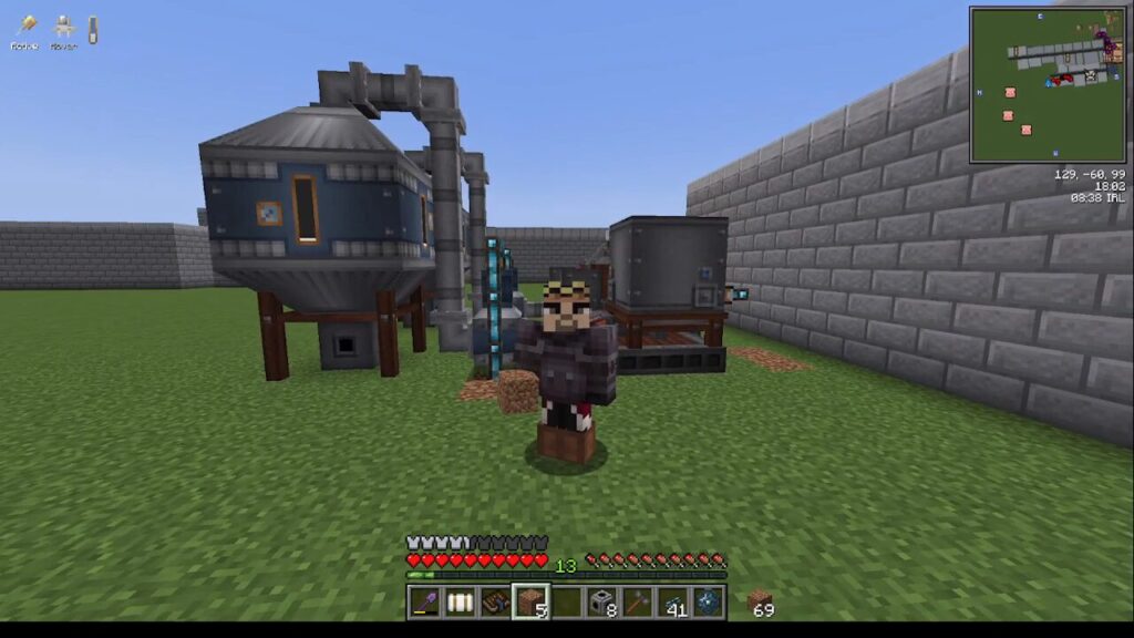 Minecraft player standing in a grassy yard beside industrial machines: a large blue processing unit on stilts connected by pipes, with a gray reactor nearby and a stone wall to the right.