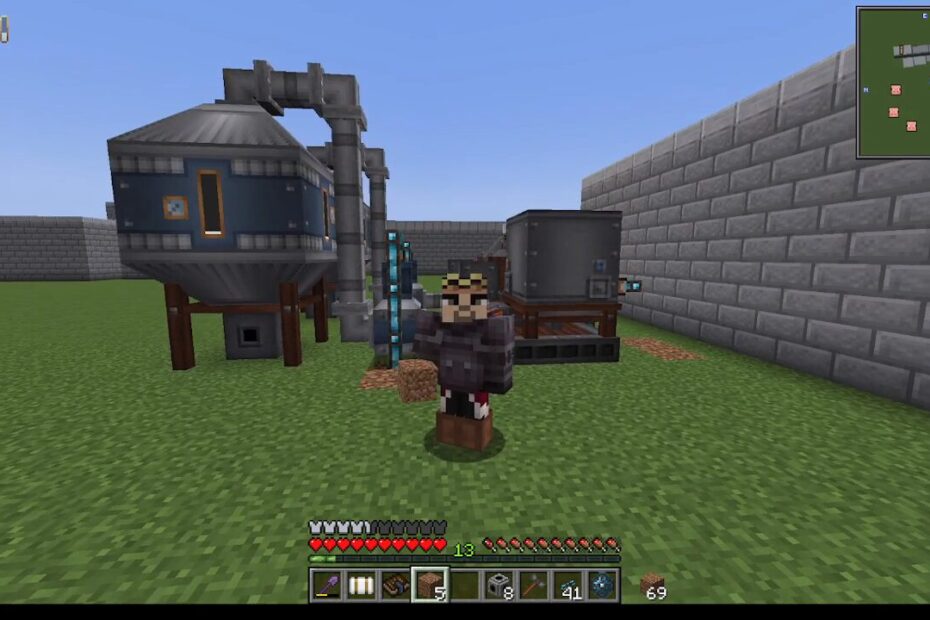 Minecraft player standing in a grassy yard beside industrial machines: a large blue processing unit on stilts connected by pipes, with a gray reactor nearby and a stone wall to the right.