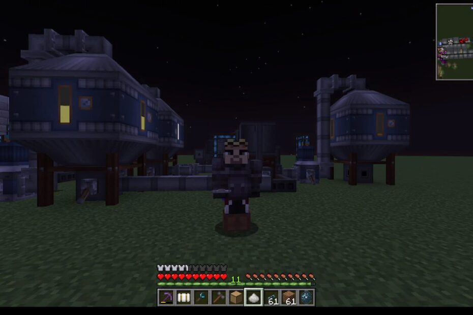 Minecraft character stands on grassy terrain at night with blue modular buildings behind, inventory and health bar visible at bottom.